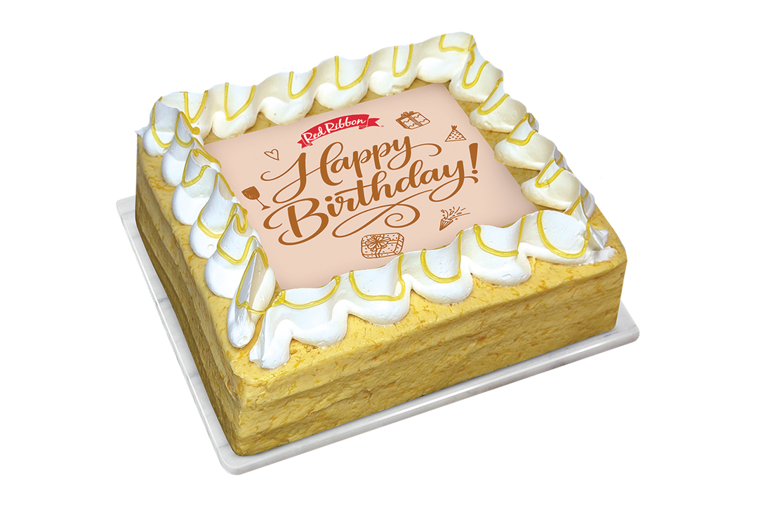 A square birthday cake from Red Ribbon Bakery, featuring a golden-yellow sponge cake base, white piped whipped cream borders, and a central peach-colored plaque with 'Happy Birthday!' written in brown script.
