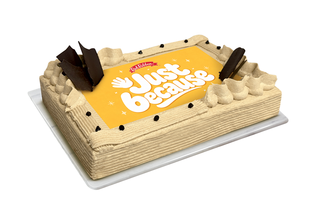 A rectangular cake with beige frosting, chocolate decorations, and a yellow top panel that reads 'Just because' with a small red 'Red Ribbon' logo, shown on a white background.