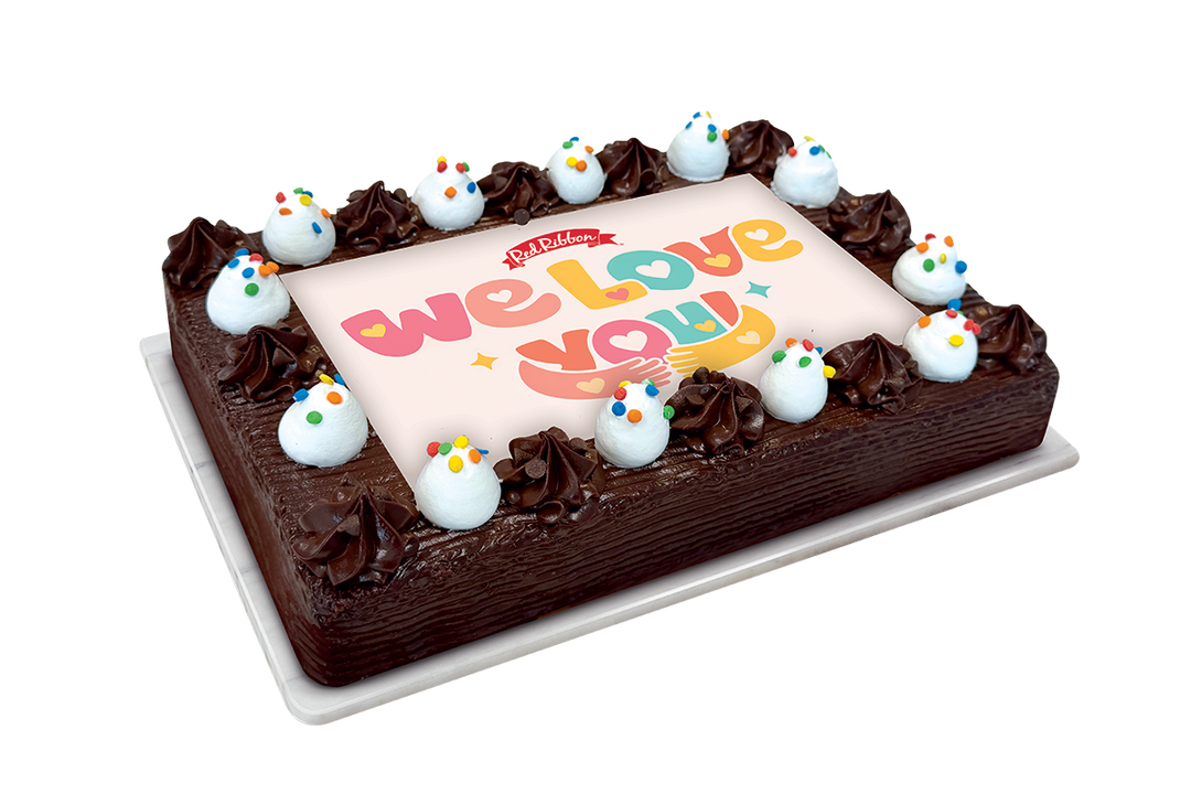 A rectangular chocolate cake on a white tray, decorated with piped chocolate frosting, white frosting dollops topped with colorful sprinkles, and a large pink icing panel that reads 'we love you' in playful multicolor lettering. The logo at the top of the lettering appears to be 'The Happy Cake'.