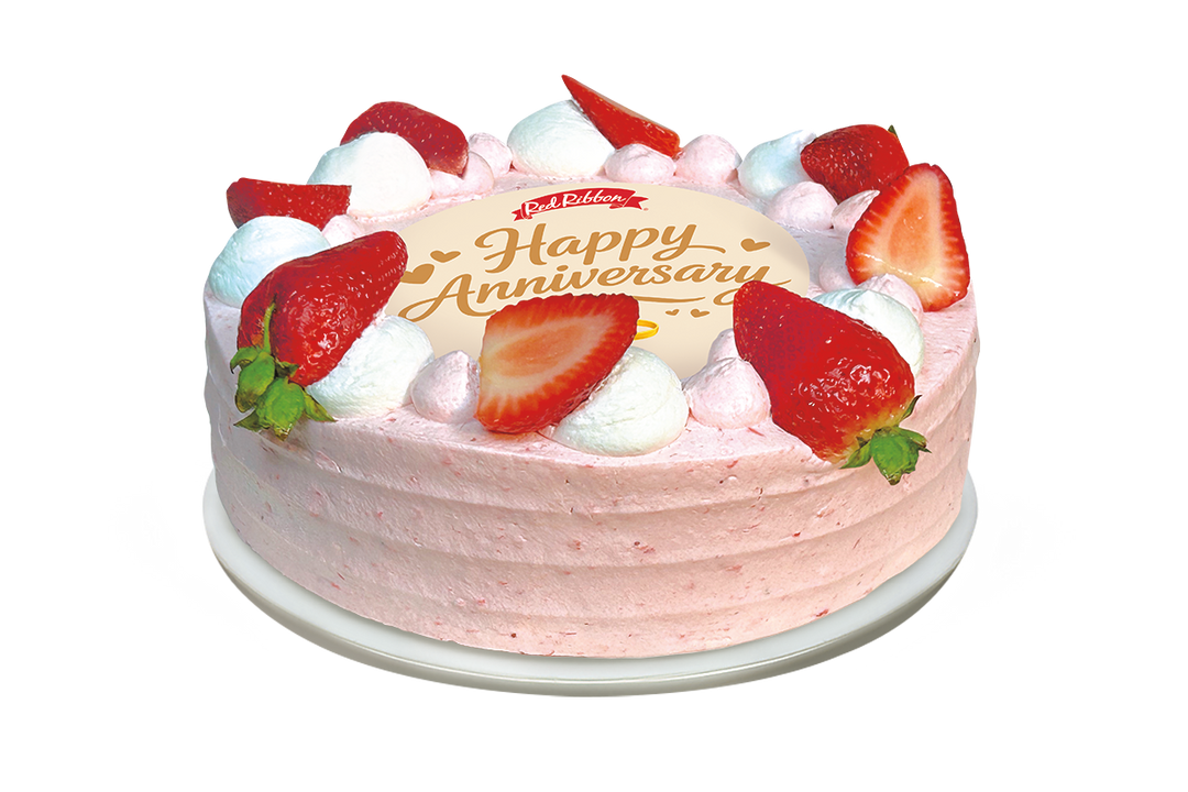 A round cake with light pink frosting and a decorative border of alternating white and pink whipped cream dollops, fresh strawberry halves, and whole strawberries. The center of the cake features a circular white topper displaying the Red Ribbon logo and the message "Happy Anniversary" in gold script, accented by small hearts and a wedding ring illustration.