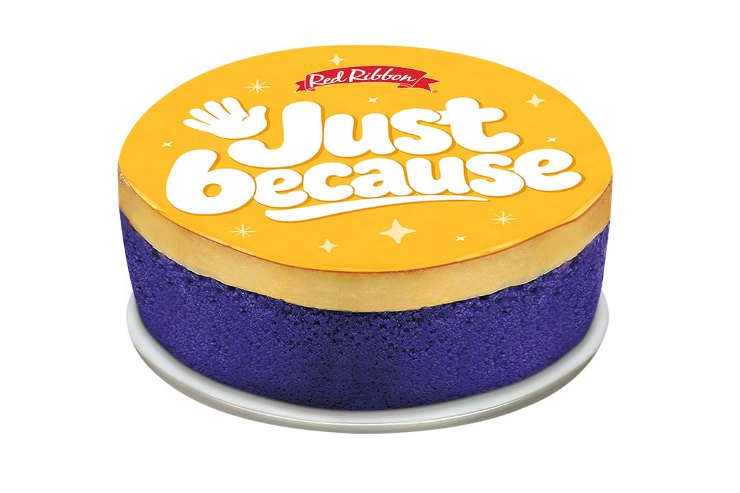 A round purple cake topped with a layer of yellow flan and a large circular yellow topper. The topper displays the Red Ribbon logo, the message "Just because" in bold white letters, and a waving hand illustration surrounded by small stars.