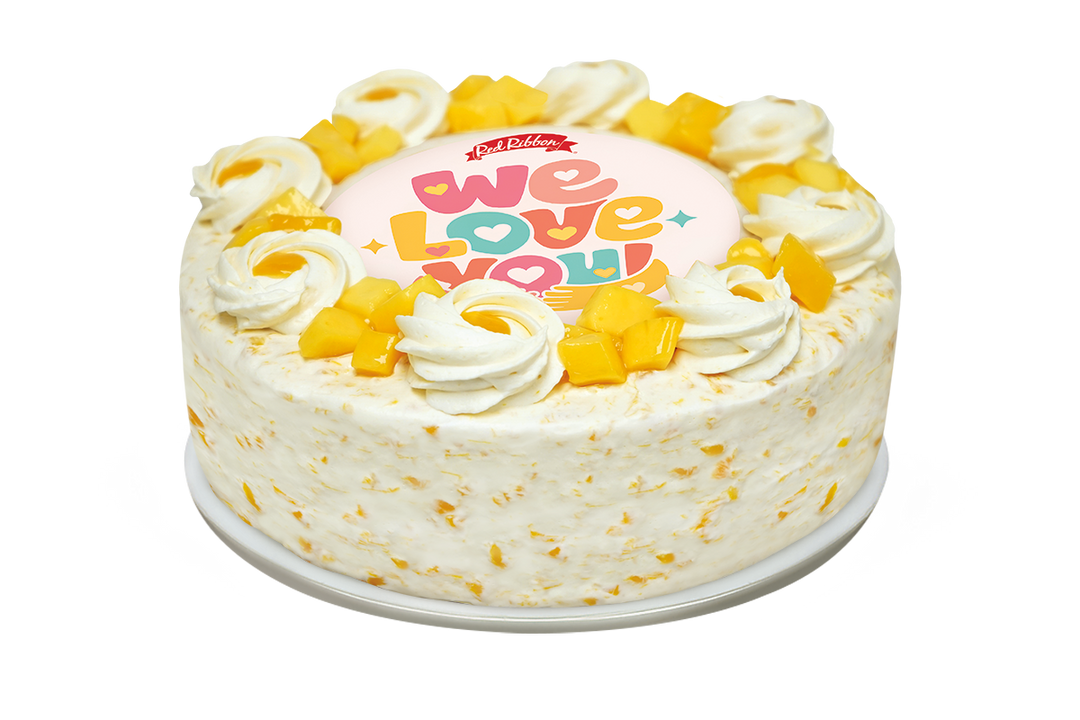 A round frosted cake with white cream and yellow mango chunks, topped with a pink topper that reads 'We Love You' in colorful letters. The cake is presented on a white background.