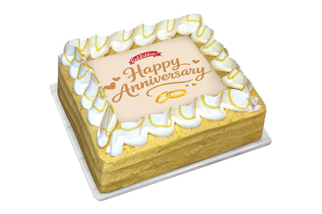 A square mango cake with a border of white piped frosting drizzled with yellow sauce. The center features a square white topper displaying the Red Ribbon logo, the message "Happy Anniversary" in gold script, and two interlocking gold wedding rings.
