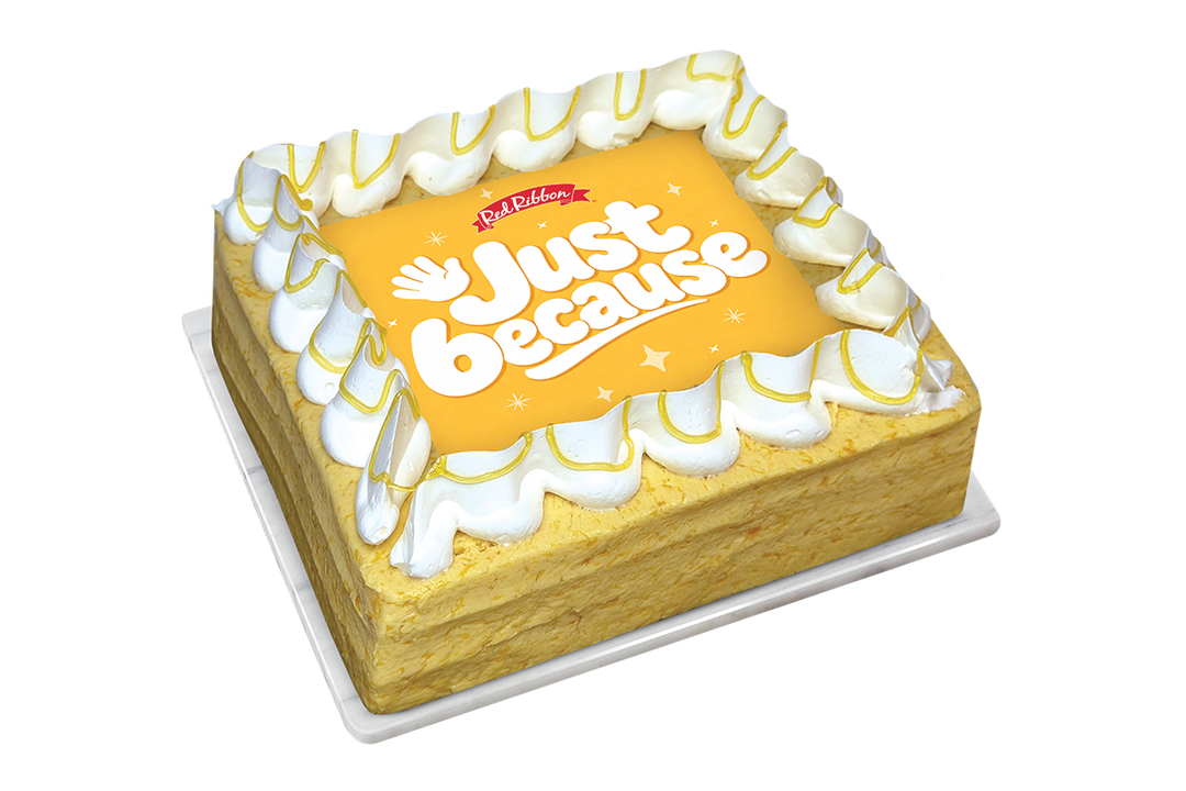 A square mango cake with a border of white piped frosting drizzled with yellow sauce. The center features a square yellow topper displaying the Red Ribbon logo, the message "Just because" in bold white letters, and a waving hand illustration.