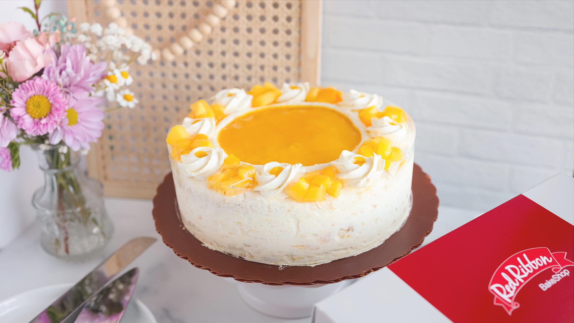 Red Ribbon Mango Supreme Cake on a cake stand with pink and white flowers.