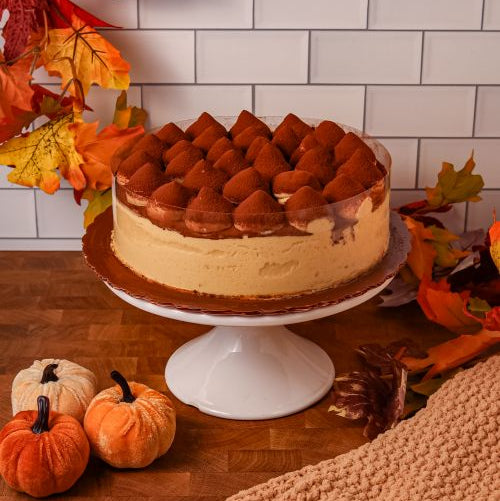 Round tiramisu cake topped with cocoa powder, displayed on a cake stand with fall decor.